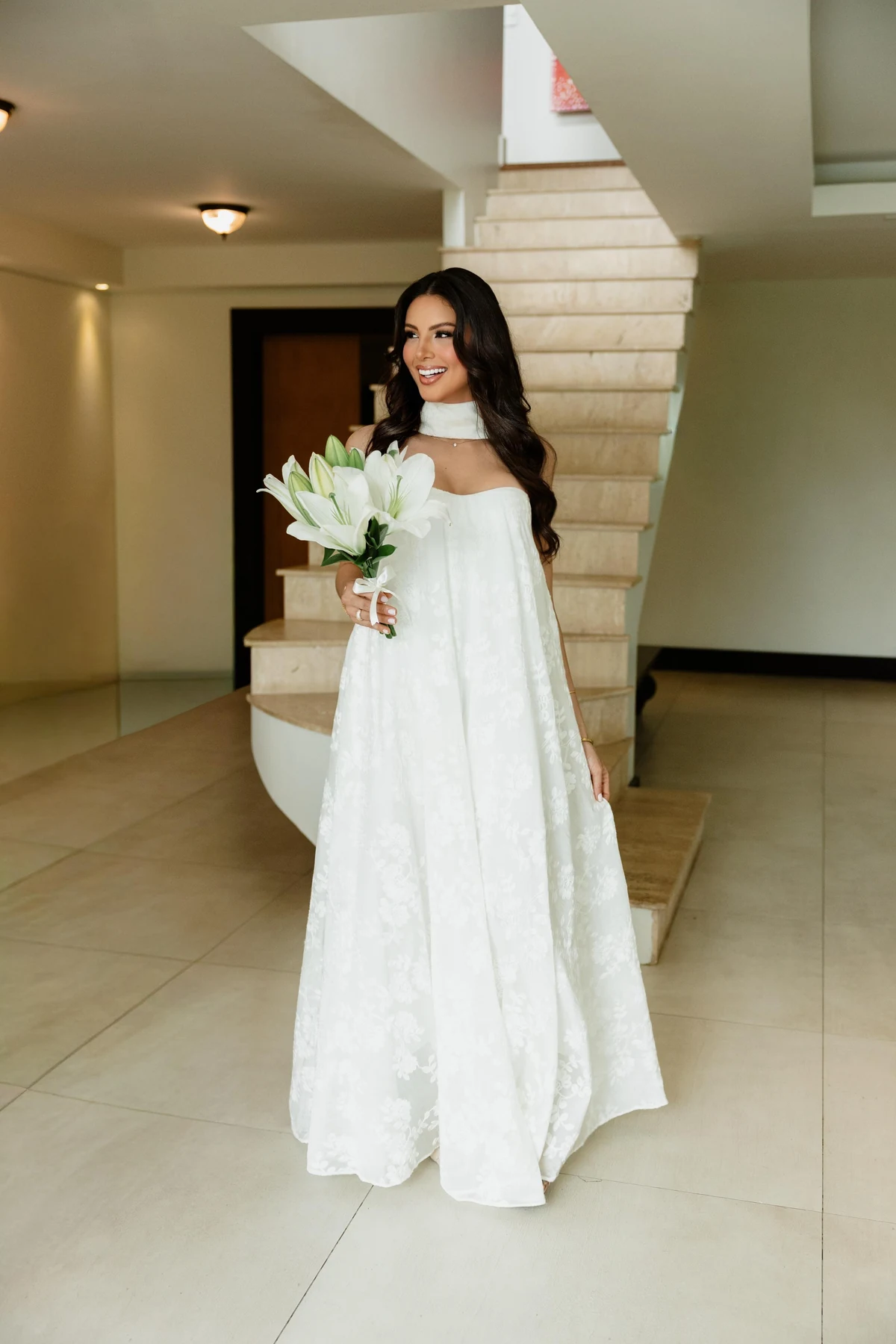 Noiva com vestido branco e buquê de lírios em ambiente interno.